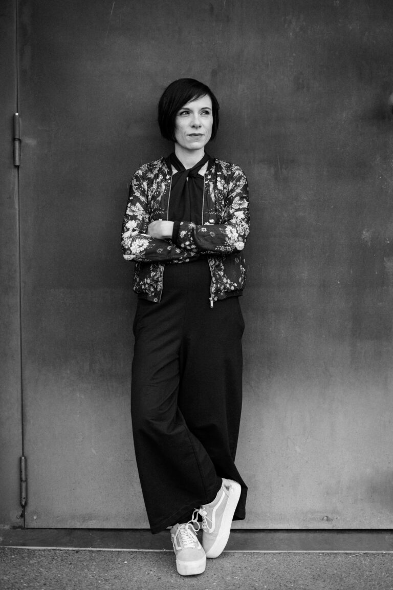 Dr. Eliane Retz steht vor einer Metalltür und lehnt mit verschränkten Armen daran an. Schwarz-weiß Foto. Branding Portraits Saarbrücken